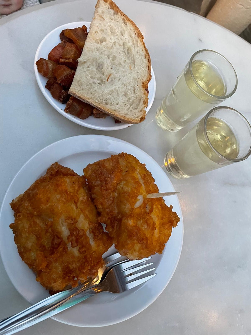 Two plates of food with fried items and bread, accompanied by two glasses of white wine on a light surface.